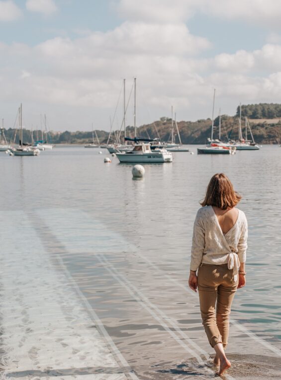 Une femme vue de dos s'avance vers l'eau de l'estuaire de la Rance en descendant un ponton à Pleurtuit, en Bretagne. Devant elle, des voiliers sont attachés à leurs bouées. Sur l'autre rive, la côte boisée de l'estuaire laisse entrevoir la rive droite de la Rance.