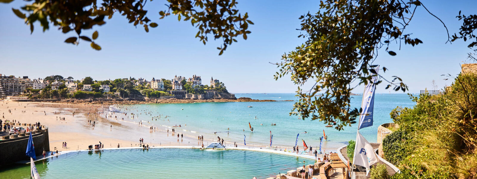 Vue en hauteur de la Piscine d'eau de mer située à Dinard au bord de la plage. De l'autre côté de la plage, les villas de la Pointe de la Malouine se trouve au dessus du sentier côtier. Sur la plage, des personnes se baignent et au bord de la piscine, le bar accueille des clients installés au soleil.