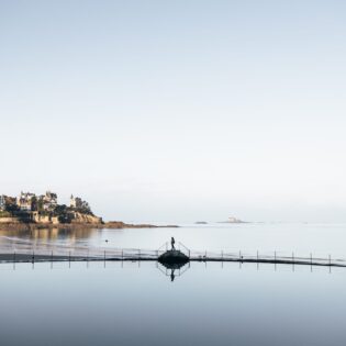 Une jeune fille marche au bord de la piscine à Dinard durant l'hiver. Derrière elle, des villas élégantes servent de toile de fond à la scène paisible. La piscine est tranquille et reflète les nuances grises du ciel hivernal.