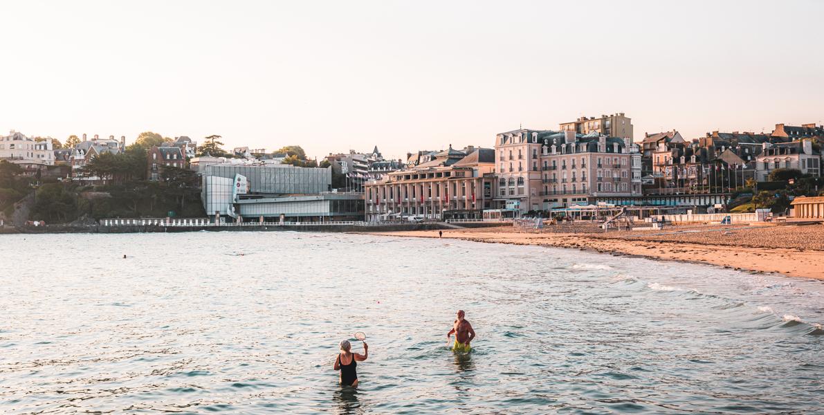 Sur la Plage de l'Écluse à Dinard, un couple de retraités se baigne dans la mer sous une lumière douce de début de journée. La lumière matinale teinte l'environnement de couleurs pastel, et le calme de la mer ajoute une sérénité à l'atmosphère. Le couple, en pleine baignade, semble apprécier ce moment paisible, entouré par la beauté naturelle de la plage au lever du soleil. Crédit photo : lezbroz