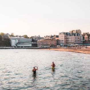 Sur la Plage de l'Écluse à Dinard, un couple de retraités se baigne dans la mer sous une lumière douce de début de journée. La lumière matinale teinte l'environnement de couleurs pastel, et le calme de la mer ajoute une sérénité à l'atmosphère. Le couple, en pleine baignade, semble apprécier ce moment paisible, entouré par la beauté naturelle de la plage au lever du soleil.
