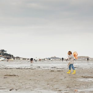 Un enfant marche sur la plage à marée basse près de Dinard, en Bretagne, France, recherche des coquillages et des crustacés dans un cadre pittoresque. L'enfant est équipé d'un seau et d'un outil de fouille, profite de cette activité traditionnelle de pêche à pied. Le ciel est légèrement nuageux, laissant filtrer la lumière douce du soleil couchant.