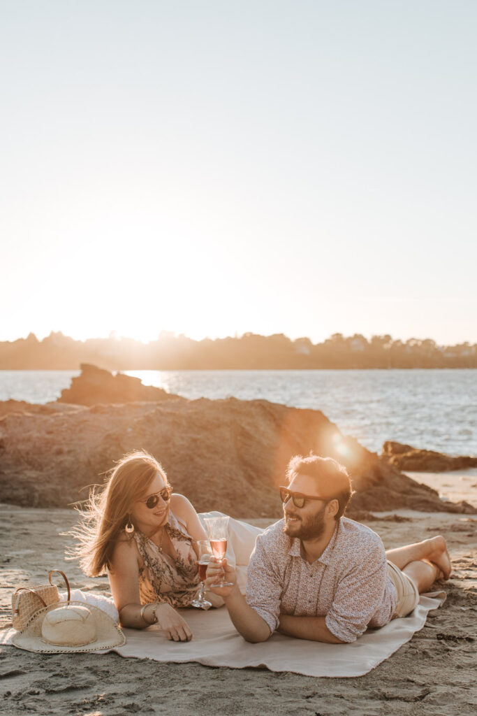 Scène romantique. Un couple allongé sur une fouta, buvant une coupe de champagne regarde le coucher du soleil à Lancieux sur la plage des Briantais