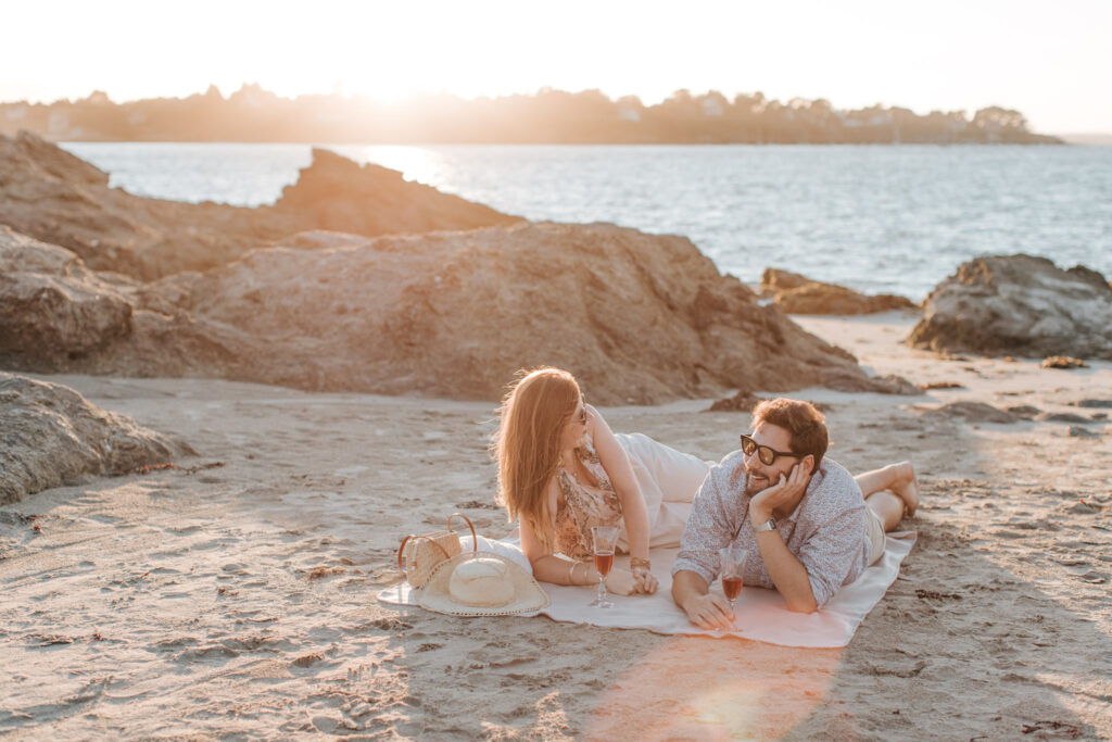 Scène romantique. Un couple allongé sur une fouta, buvant une coupe de champagne regarde le coucher du soleil à Lancieux sur la plage des Briantais