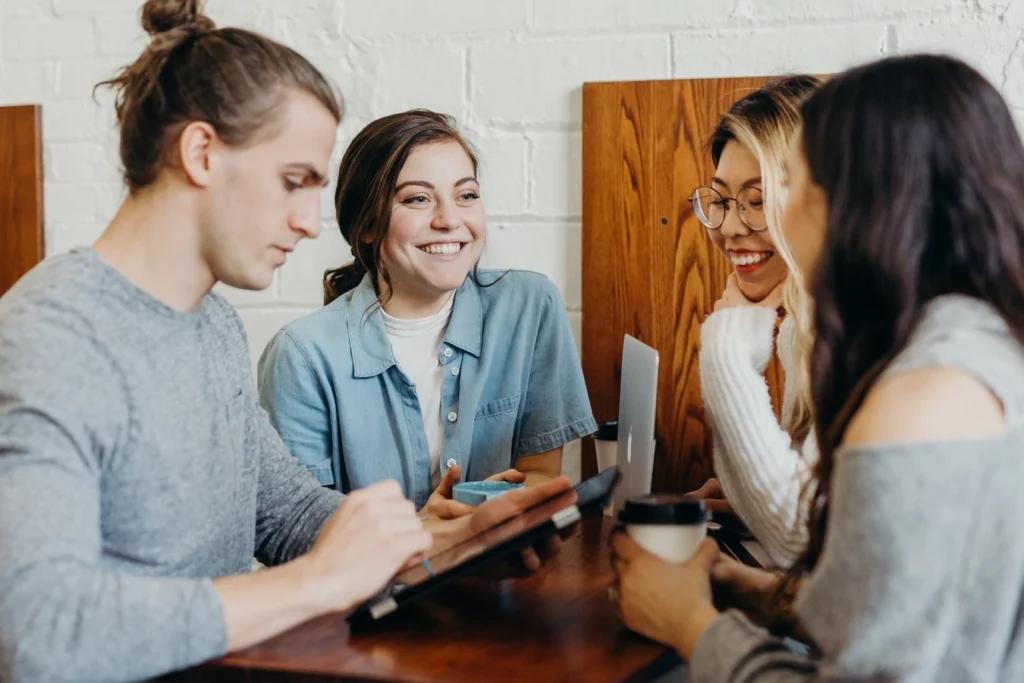 Photo de groupe de quatre professionnels souriants, debout dans un bureau lumineux. Trois femmes et un homme, représentant une diversité d'âges et de races, discutent autour d'une table avec des documents et des ordinateurs portables. Ils semblent engagés dans une collaboration fructueuse. espace pro partenariat hébergements collectif