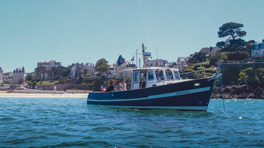 Le bateau de L'échappée Malouine navigue devant la Grande Plage de Saint-Lunaire et la Pointe du Décollé. Privatisez une croisière pour vos amis et votre famille sur la Côte d'Émeraude, pour découvrir la Baie de Saint-Malo en bateau !