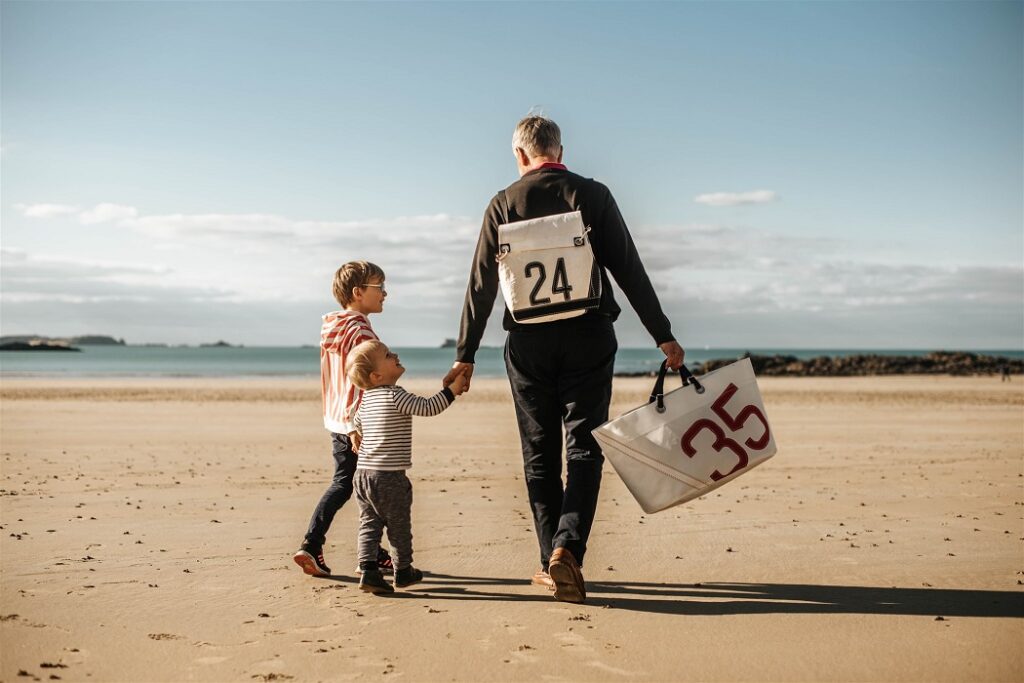 Vent De Voyage - un père et ses fils se baladent sur la plage avec des sacs de voyage faits à partir de voiles de bateaux.