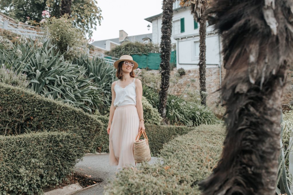 Une femme habillée de blanc avec un chapeau de paille se promène à Dinard entre les buissons et la végétation méditerranéenne.