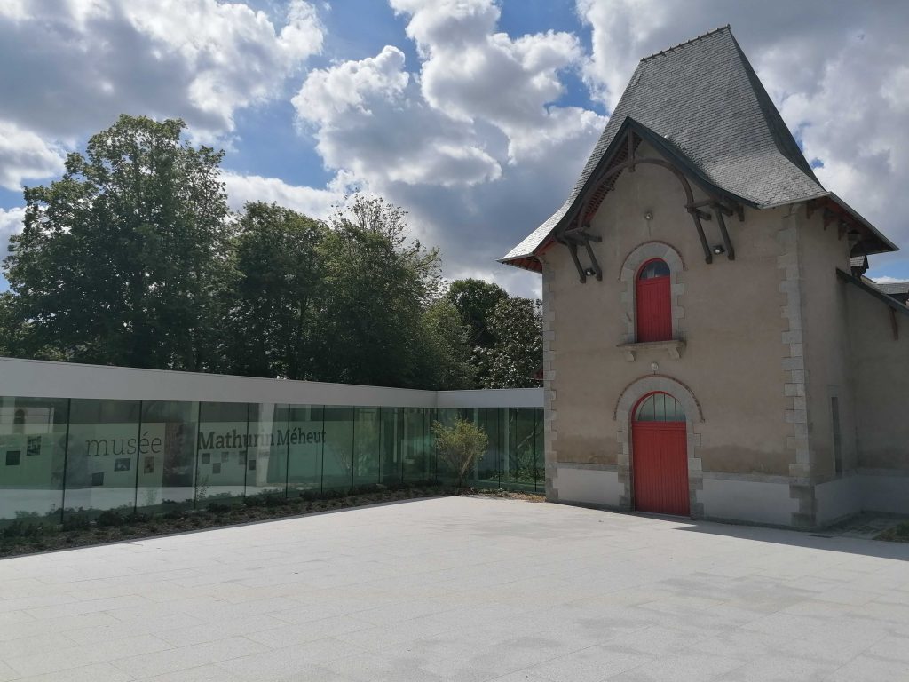 Entrée du Musée Mathurin Méheut à Lamballe composé d'une structure en verre sur la gauche et d'une bâtisse ancienne à un étage aux portes et fenêtres rouges.