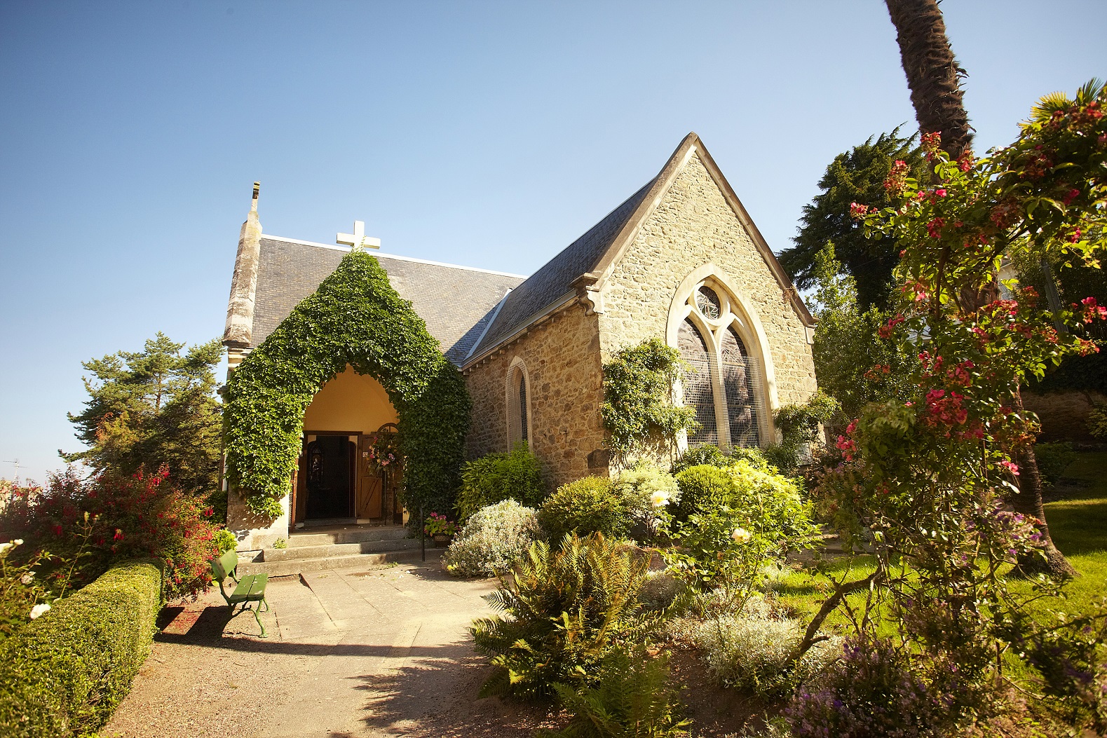 Façade en pierres de l'Eglise Anglicane St Barts De Dinard. Un chemin d'accès entouré de verdure mène à la porte d'entrée en forme d'arche.