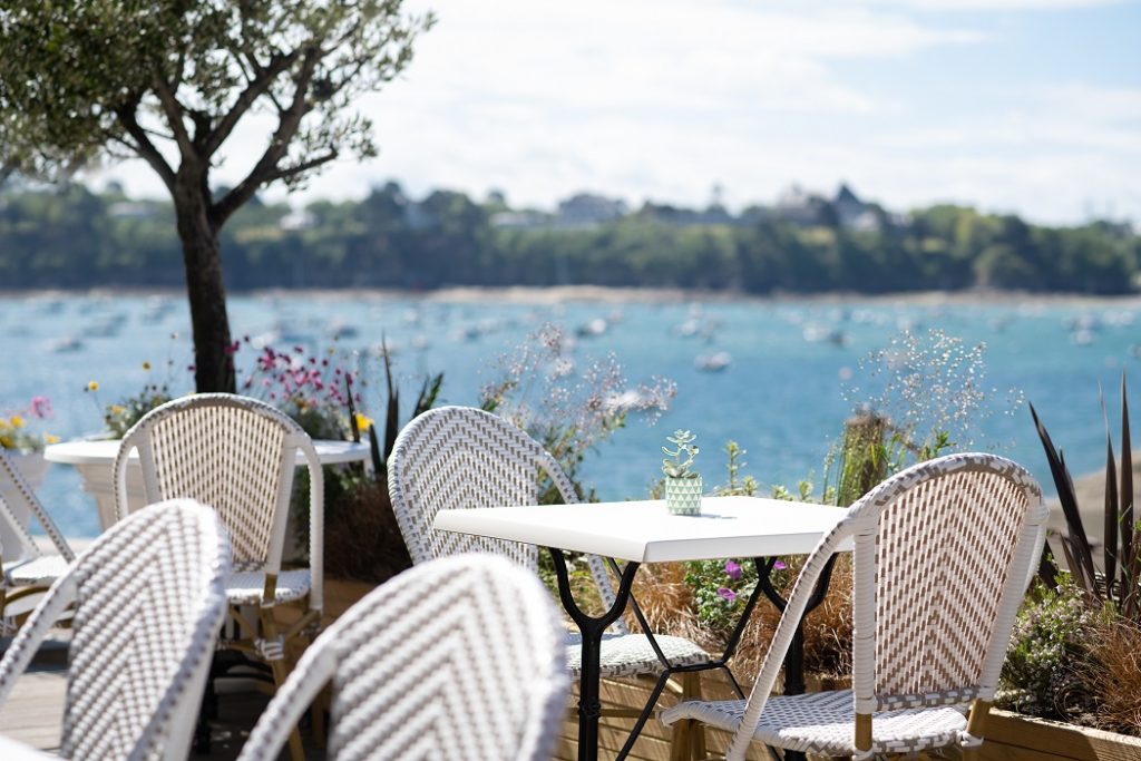 terrasse en bord de mer du restaurant La Vallée à Dinard, avec des chaises en osier tissées de blanc et de beige, disposées autour de tables blanches. En arrière-plan, on aperçoit la baie du Prieuré calme, avec des bateaux ancrés et une côte verdoyante au loin.