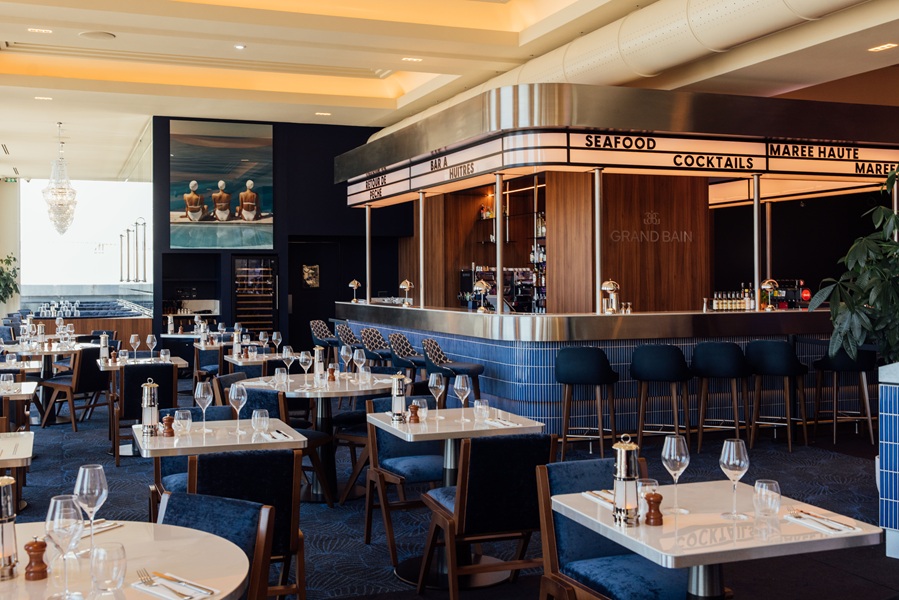 Photo de la grande salle du restaurant Le Grand Bain du Casino Barrière de Dinard, élégante, lumineuse et soigneusement dressée, avec des tables couvertes de verres et de couverts. Au fond, un bar moderne aux tons bleu et bois affiche des enseignes comme « Seafood » et « Cocktails », avec des tabourets alignés. L’ambiance est raffinée, ponctuée de détails décoratifs comme un grand tableau représentant trois nageurs et un lustre en cristal. Crédit Agence Pancake