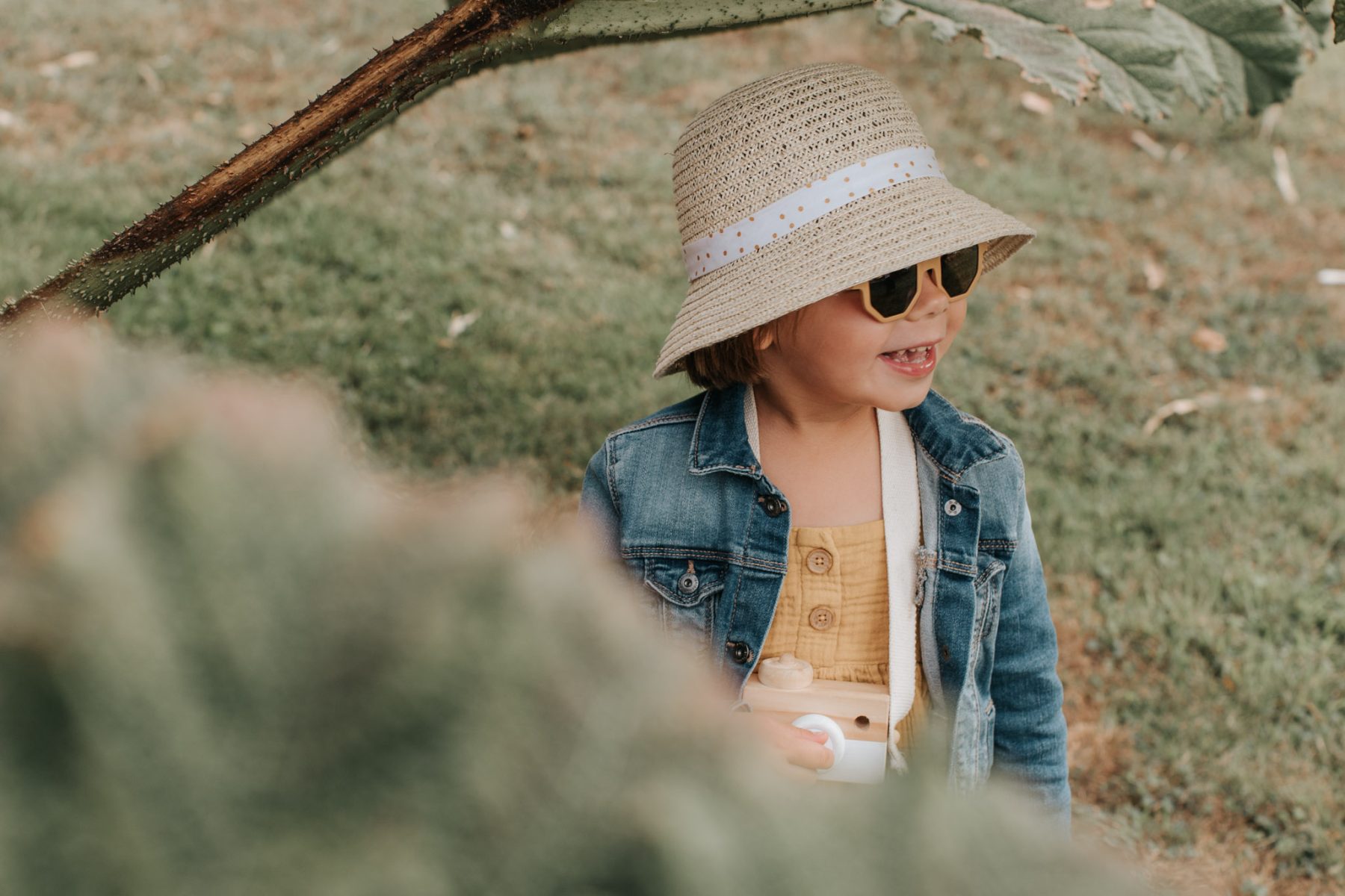 Petite fille portant un chapeau de paille, des lunettes de soleil et une veste en jean, debout dans l’herbe sous une branche d’arbre dans le jardin de Montmarin à Pleurtuit