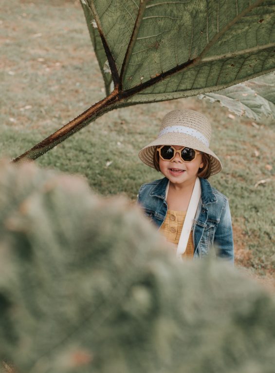 Petite fille portant un chapeau de paille, des lunettes de soleil et une veste en jean, debout dans l’herbe sous une branche d’arbre dans le jardin de Montmarin à Pleurtuit